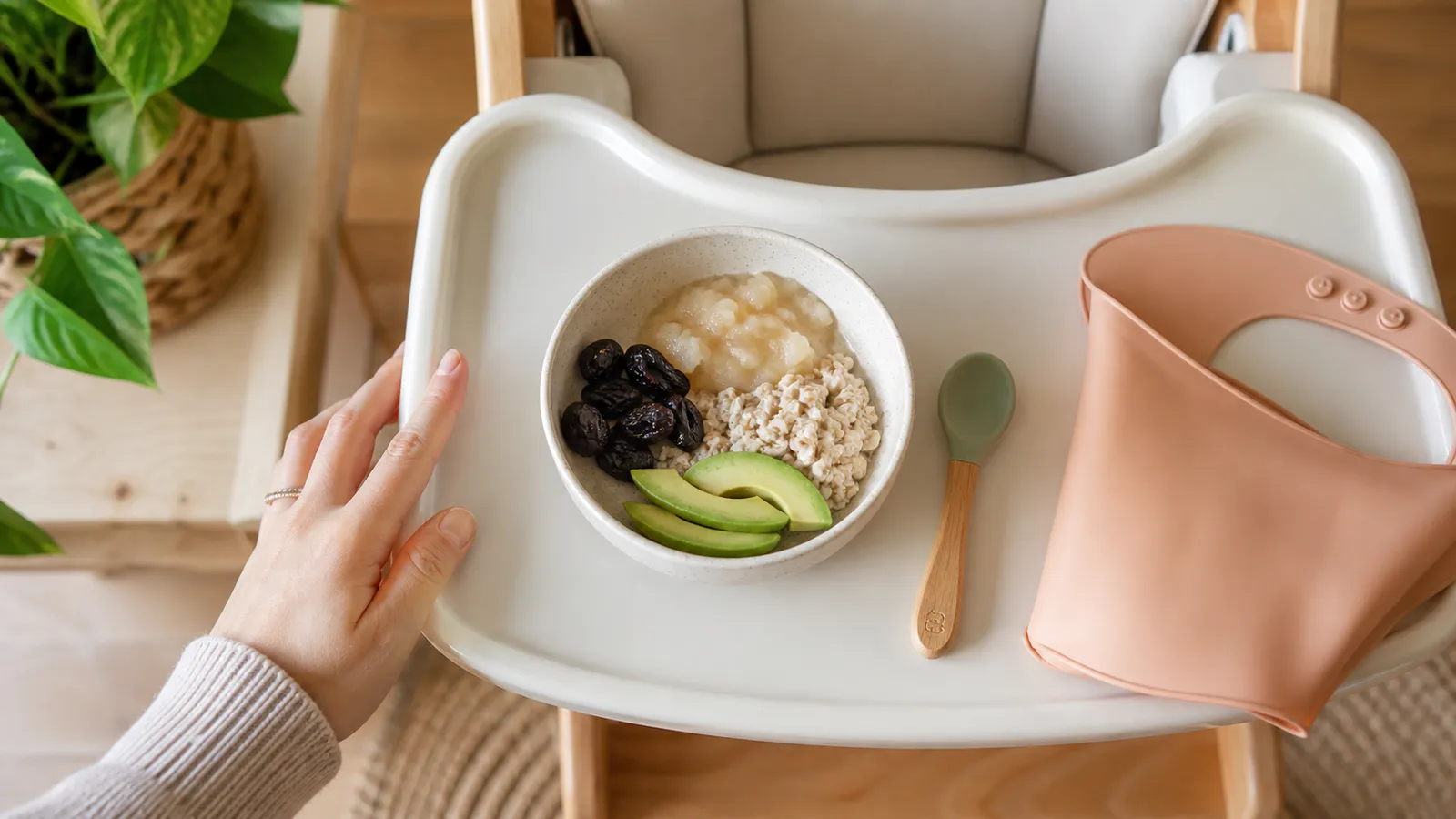 Overhead view of a high chair tray with a bowl of pear, prunes, oats, avocado, a baby spoon, and a soft silicone bib.